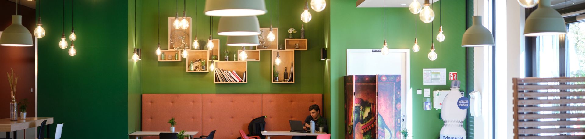 Es wird ein Teilausschnitt des FS-Dailys gezeigt. Es sind Stühle, Tische und viele Leuchten an der Decke zu sehen. Die Wand strahlt grün.